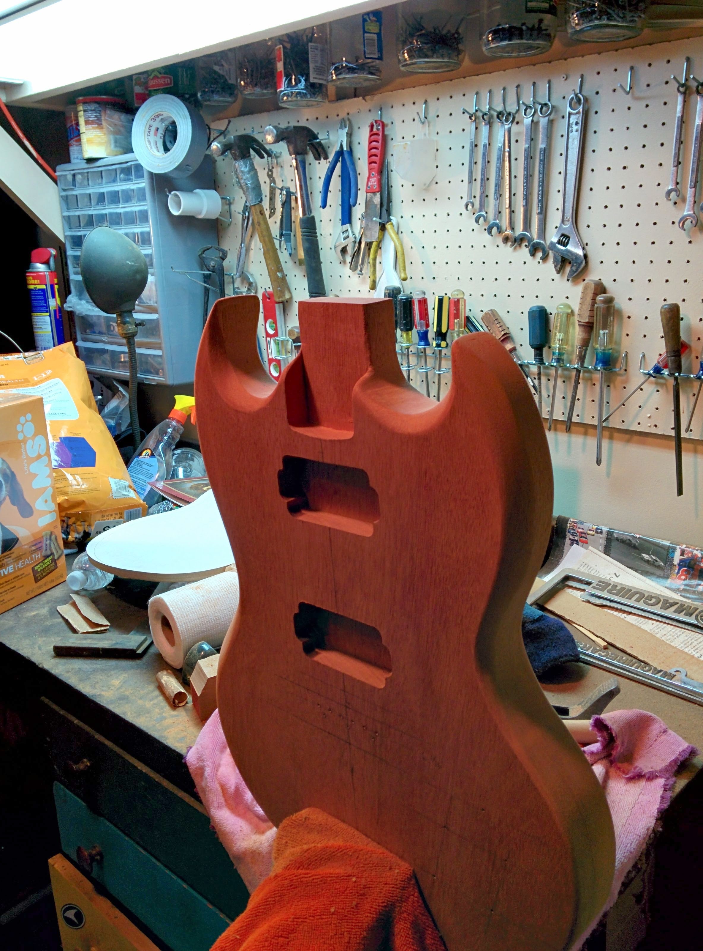 Mahogany SG-style guitar body in progress on a workbench, with pegboard of tools behind